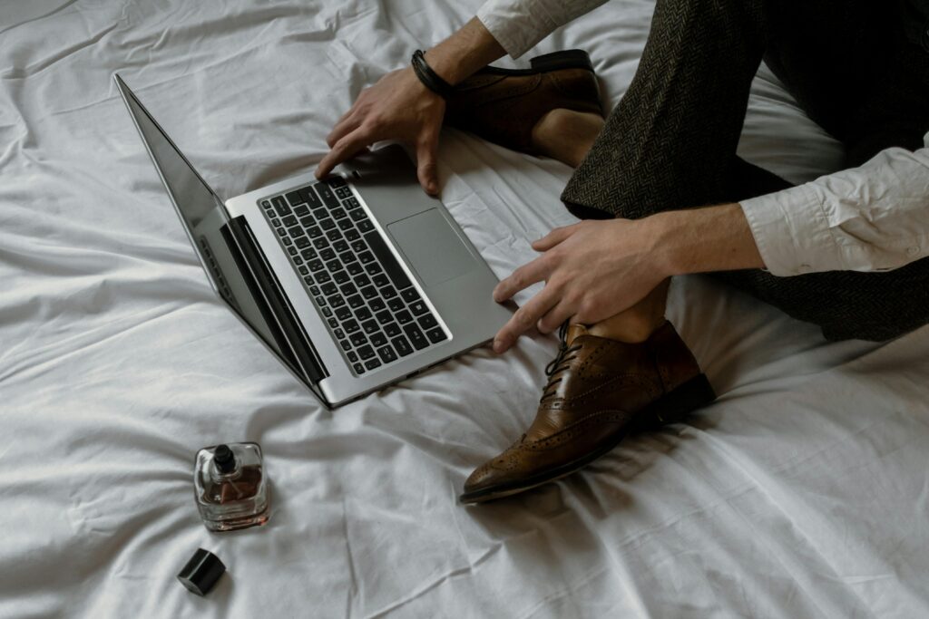 Adult man in formal attire working on a laptop while sitting on bed indoors.